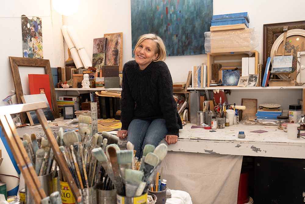 Visual artist Krista Machovina sits on a table in an art studio wearing a black shirt and blue jeans